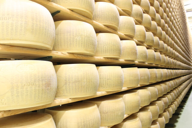 Auf dem Bild sind viele Käselaibe, wahrscheinlich Parmigiano-Reggiano, aufgestapelt und gelagert zu sehen.