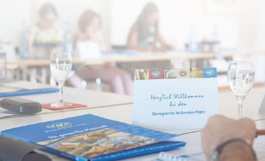 Auf dem Bild ist eine Willkommenskarte für ein Treffen der Ökologischen Molkerei Allgäu zu sehen, während Teilnehmer im Hintergrund an einem Tisch sitzen.