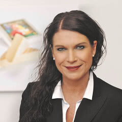 Das Bild zeigt eine Frau mit langen, dunklen Haaren, die in formeller Kleidung lächelt, vor einem hellen Hintergrund mit Bildern.