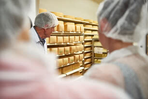 Auf dem Bild sind mehrere Personen zu sehen, die in einem Lagerraum mit Regalen voller Käselaibe stehen.