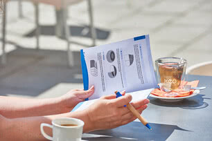 Auf dem Bild sind eine Person, die ein Dokument mit Anleitungen oder Informationen in der Hand hält, sowie eine Tasse Kaffee und ein Glas Tee zu sehen.