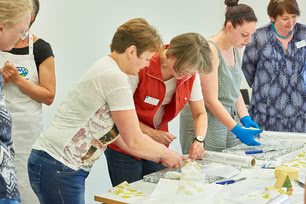 Auf dem Bild sind mehrere Personen zu sehen, die gemeinsam an einem Tisch an einem Projekt arbeiten, wahrscheinlich im Bereich von Handwerk oder Kunst.