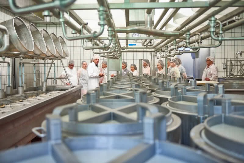 Auf dem Bild ist eine Gruppe von Menschen in einem Produktionsraum zu sehen, die an Maschinen und Behältern stehen und wahrscheinlich eine Schulung oder Besichtigung in einer Lebensmittelverarbeitungseinrichtung durchführen.