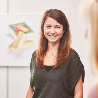 Auf dem Bild ist eine lächelnde Frau zu sehen, die in einem hellen Raum steht, während im Hintergrund ein Bild eines Snacks oder einer Süßigkeit zu erkennen ist.