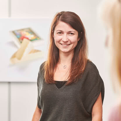 Auf dem Bild ist eine lächelnde Frau zu sehen, die in einem hellen Raum steht, während im Hintergrund ein Bild eines Snacks oder einer Süßigkeit zu erkennen ist.