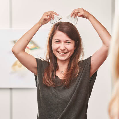 Auf dem Bild ist eine lächelnde Frau zu sehen, die mit beiden Händen zwei weiße Gegenstände über ihrem Kopf hält, die wie Ohren aussehen.