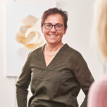 Auf dem Bild ist eine lächelnde Frau in einem grünen Oberteil zu sehen, die auf eine andere Person blickt.