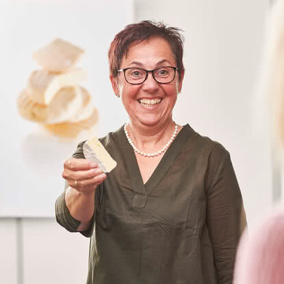 Auf dem Bild ist eine lachende Frau zu sehen, die ein Stück Käse in der Hand hält.