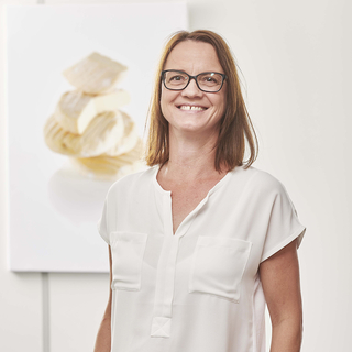 Auf dem Bild ist eine Frau mit Brille zu sehen, die lächelt, während im Hintergrund eine Bildergalerie mit Käse abgebildet ist.
