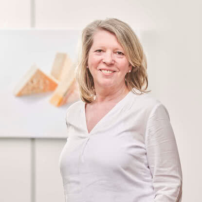 Auf dem Bild ist eine Frau in einem weißen Oberteil zu sehen, die lächelt, während im Hintergrund ein Kunstwerk mit Käsestücken abgebildet ist.