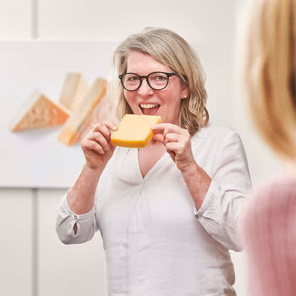 Auf dem Bild ist eine Frau zu sehen, die ein Stück Käse hält und lächelt.