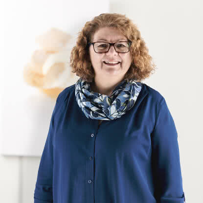 Auf dem Bild ist eine lächelnde Person mit lockigem, braunen Haar und Brille zu sehen, die ein blaues Hemd und einen gemusterten Schal trägt.