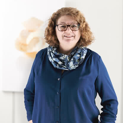 Auf dem Bild ist eine lächelnde Person mit lockigem Haar und Brille zu sehen, die ein dunkelblaues Hemd und einen gemusterten Schal trägt.