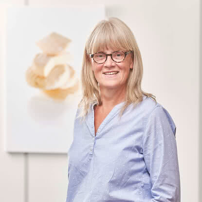 Auf dem Bild ist eine lächelnde Frau mit blonden Haaren und einer Brille zu sehen, die sich vor einem unscharfen Kunstwerk befindet.