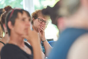 Auf dem Bild sind mehrere Personen in einer Gesprächssituation zu sehen, wobei eine Frau im Vordergrund nachdenklich wirkt.
