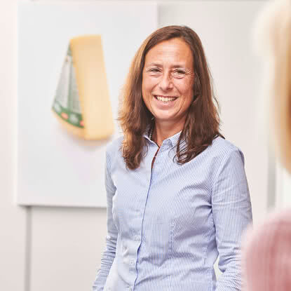 Auf dem Bild ist eine lächelnde Frau in einem gestreiften Hemd zu sehen, die vor einem Kunstwerk steht, das einen Käsestück darstellt.