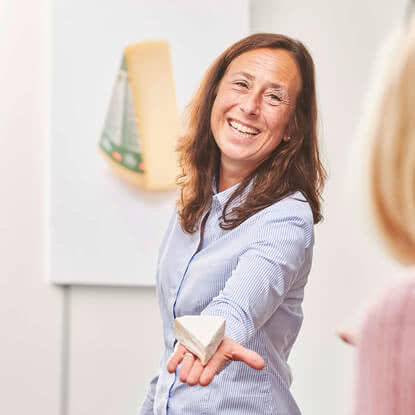 Auf dem Bild ist eine lächelnde Frau zu sehen, die einem anderen Menschen ein Stück Käse anbietet.