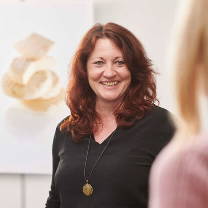 Auf dem Bild ist eine lächelnde Frau mit roten Haaren zu sehen, die in einem hellen Raum posiert. Im Hintergrund sind unscharfe Objekte zu erkennen.