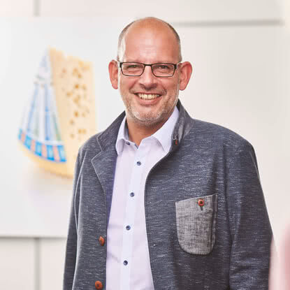 Auf dem Bild ist ein Mann zu sehen, der lächelt und einen grauen Blazer trägt, im Hintergrund ist ein Käse zu erkennen.