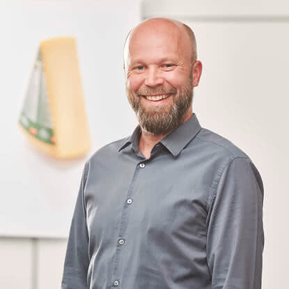 Auf dem Bild ist ein lächelnder Mann mit Bart zu sehen, der ein graues Hemd trägt, vor einem hellen Hintergrund mit einem Käse.