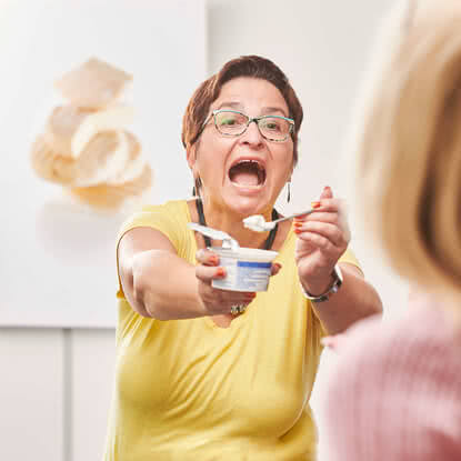 Auf dem Bild ist eine Frau zu sehen, die begeistert einen Löffel aus einem Becher reicht, während sie offensichtlich etwas mitteilen oder anbieten möchte.