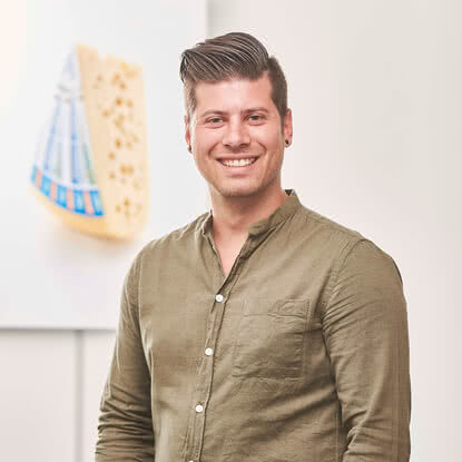 Auf dem Bild ist ein lächelnder Mann in einem grünen Hemd zu sehen, der vor einem Kunstwerk mit einem Stück Käse im Hintergrund steht.