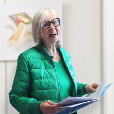Auf dem Bild ist eine lächelnde Frau mit grauen Haaren und Brille zu sehen, die ein grünes Outfit trägt und ein Heft in der Hand hält.