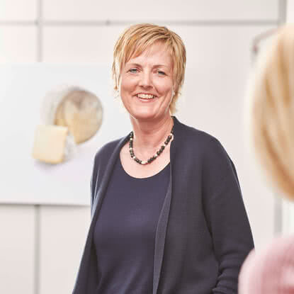 Auf dem Bild ist eine lächelnde Frau zu sehen, die in einem hellen Raum steht, während im Hintergrund Käse dargestellt ist.