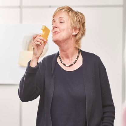 Auf dem Bild ist eine Frau zu sehen, die genießerisch ein Stück Käse hochhält und es anscheinend riecht oder betrachtet.