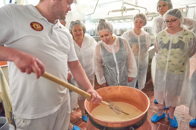 Auf dem Bild ist eine Gruppe von Personen zu sehen, die in einem Käselabor einem Fachmann beim Käsemachen zuschaut.