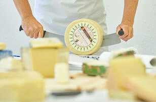 Auf dem Bild ist eine Person zu sehen, die mit einem großen Messer einen runden Käse schneidet.