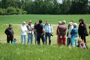 Auf dem Bild ist eine Gruppe von Menschen zu sehen, die im Freien zusammenstehen und einem Mann zuhören, der anscheinend spricht oder erläutert.