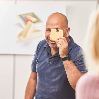 Auf dem Bild ist ein lächelnder Mann zu sehen, der ein Stück Käse vor sein Gesicht hält.