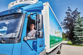 Auf dem Bild ist ein lächelnder Mann im Fahrerhaus eines blauen Lastwagens zu sehen.