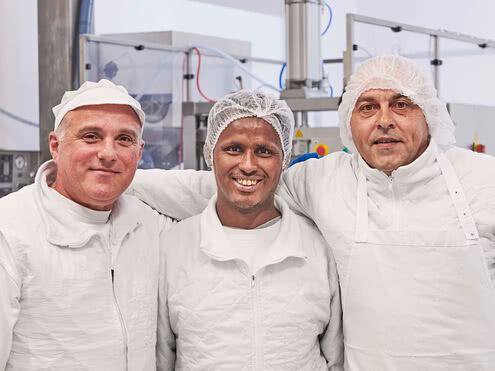 Auf dem Bild sind drei Männer in weißen Arbeitsanzügen und Haarnetzen zu sehen, die lächelnd in einer Umgebung stehen, die wahrscheinlich zu einer Lebensmittel- oder Produktionsstätte gehört.