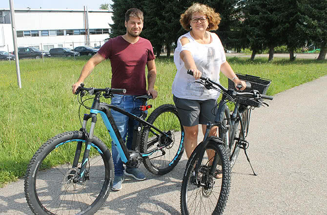 Auf dem Bild sind zwei Personen zu sehen, die neben ihren Fahrrädern stehen und lächeln.