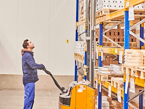 Auf dem Bild ist ein Mann zu sehen, der mit einem elektrischen Gabelstapler in einem Lager arbeitet und nach oben schaut, vermutlich um eine Palette zu erreichen.
