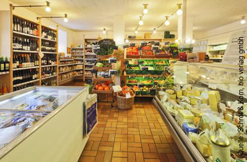 Das Bild zeigt ein gut sortiertes Lebensmittelgeschäft mit Regalen voller Flaschen, Obst, Gemüse und Käse.