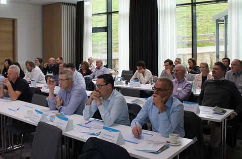 Auf dem Bild sind Teilnehmer eines Seminars oder einer Konferenz zu sehen, die aufmerksam zuhören und Notizen machen.