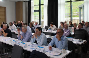 Auf dem Bild sind Teilnehmer eines Seminars oder einer Konferenz zu sehen, die aufmerksam zuhören und Notizen machen.