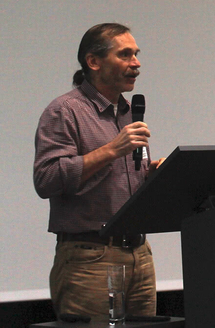 Auf dem Bild ist ein Mann zu sehen, der mit einem Mikrofon spricht, während er an einem Rednerpult steht.