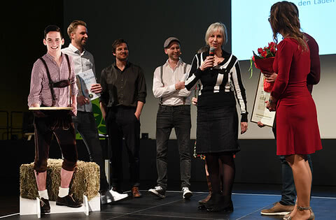 Auf dem Bild sind mehrere Personen auf einer Bühne während einer Veranstaltung zu sehen, bei der eine Frau mit Blumen einen Preis überreicht, während ein Mann in traditioneller Tracht daneben steht.