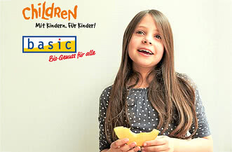 Auf dem Bild ist ein Mädchen zu sehen, das lächelt und ein Stück Obst hält, begleitet von Texten, die auf ein Kinderprojekt und biologische Genussmittel hinweisen.