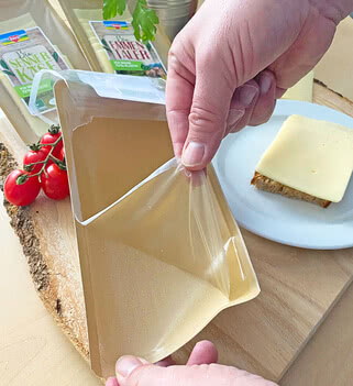 Auf dem Bild ist zu sehen, wie jemand eine Verpackung öffnet, in der wahrscheinlich Käse aufbewahrt ist, während daneben eine Scheibe Brot mit Käse liegt und frische Tomaten zu sehen sind.