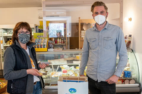 Auf dem Bild sind eine Frau und ein Mann in einem Geschäft zu sehen, wobei die Frau ein Lebensmittelprodukt in der Hand hält und der Mann eine Tüte mit Einkäufen. Beide tragen Masken.