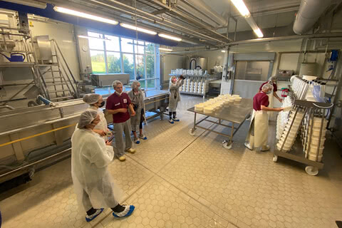 Auf dem Bild ist eine Gruppe von Personen in einem Lebensmittelbetrieb zu sehen, die in einem Produktionsraum für Käse stehen und an einem Rundgang oder einer Besichtigung teilnehmen.