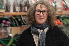 Auf dem Bild ist eine Frau mit lockigem Haar, Brille und einem Schal zu sehen, die in einem Markt oder Geschäft für frische Lebensmittel steht.