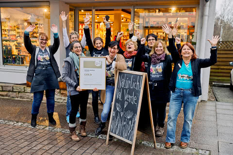 Auf dem Bild ist eine Gruppe von Frauen zu sehen, die fröhlich mit erhobenen Händen vor einem Geschäft stehen und eine Auszeichnung halten.