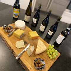 Auf dem Bild ist eine Käseplatte mit verschiedenen Käsesorten, Oliven und Trauben sowie mehreren Weinflaschen zu sehen.