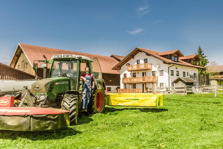 Auf dem Bild ist ein Landwirt zu sehen, der neben einem grünen Traktor und einem gelben Mähgerät auf einem Bauernhof steht, hinter ihm ist ein Wohngebäude sichtbar.